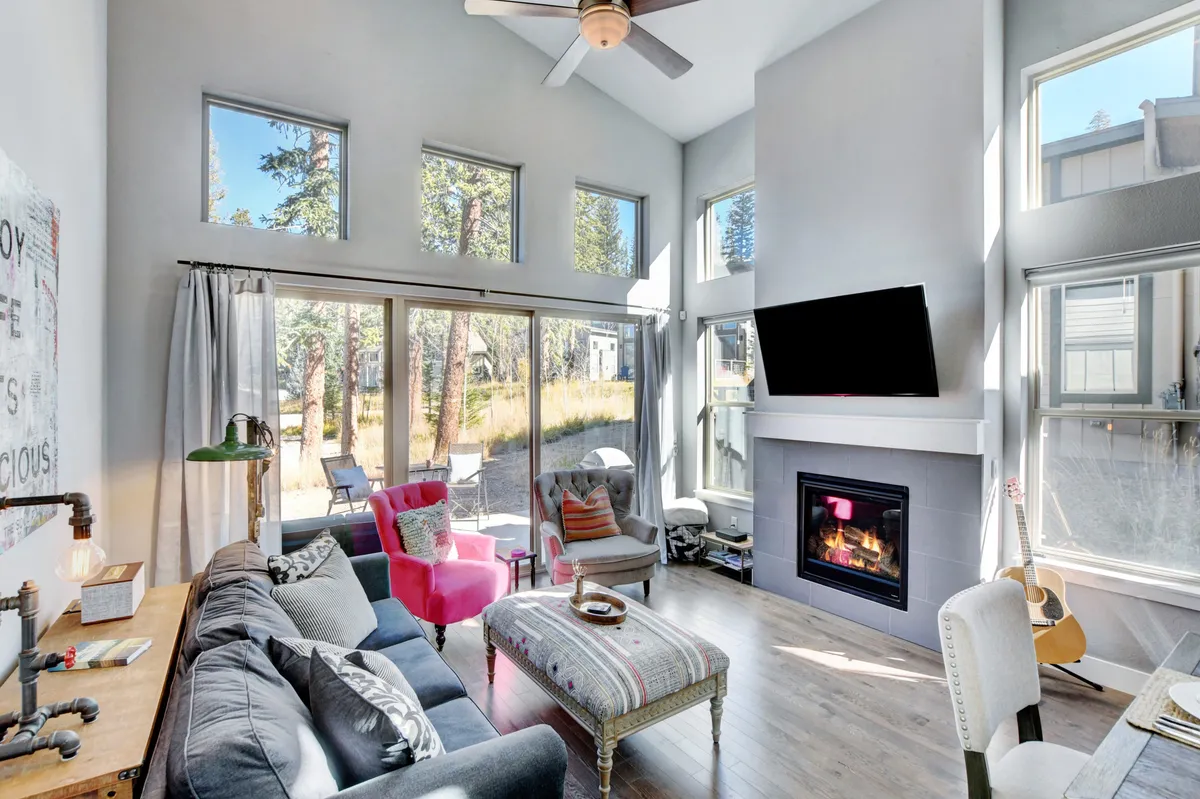A warm, open living area featuring a modern fireplace as the focal point, flanked by a comfortable sofa and a colorful accent chair. Expansive windows let in abundant natural light and frame tranquil views outside, enhancing the room's connection to the natural mountain surroundings. The space exudes both style and comfort, ideal for relaxing or socializing with family and friends.