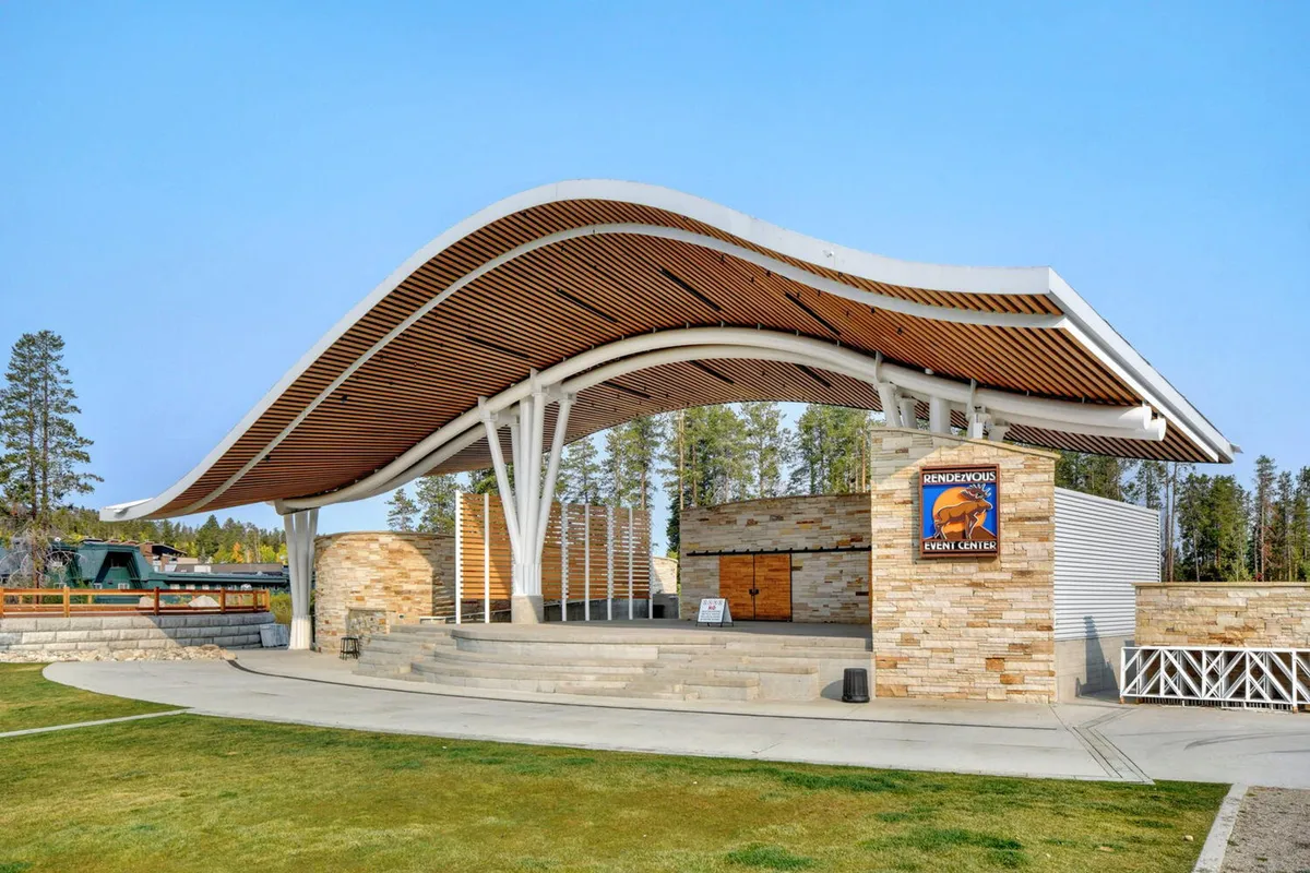 This exterior shot captures a modern event center with a unique wave-like roof structure and a welcoming stone wall. The entrance area is designed for gatherings and community events, set against the backdrop of the mountains.