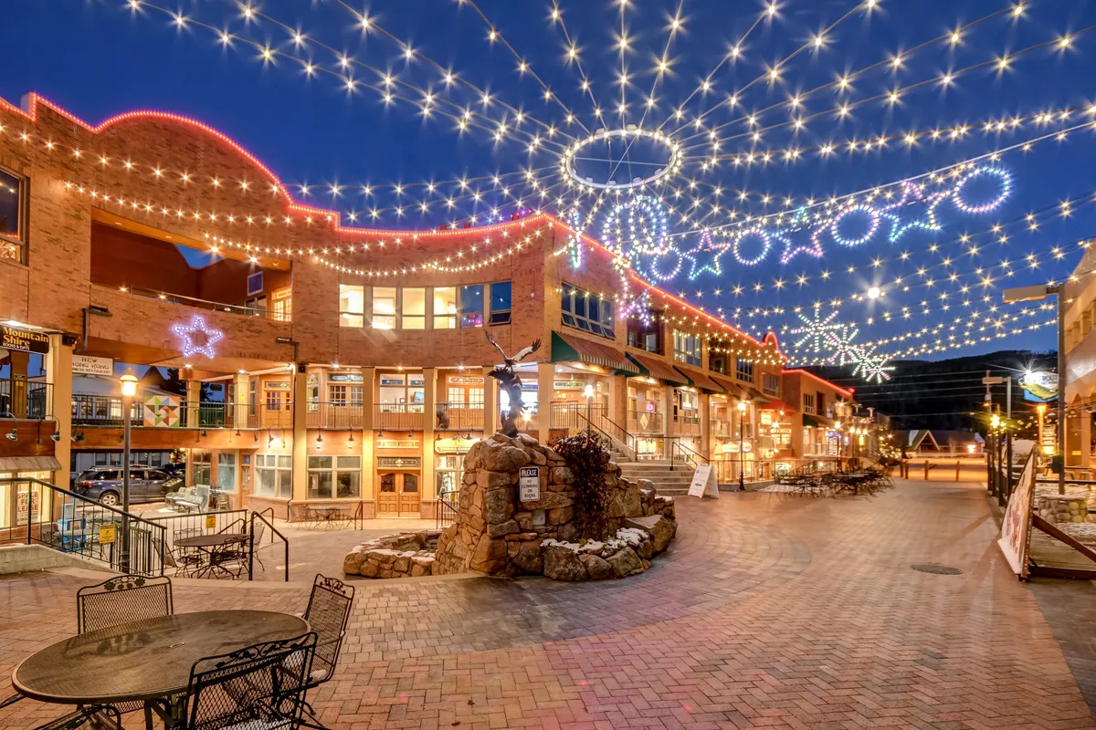 A lively exterior scene featuring string lights, decorative stars, a central sculpture fountain, and a brick pathway. The setting suggests a vibrant community space ideal for socializing or evening strolls.