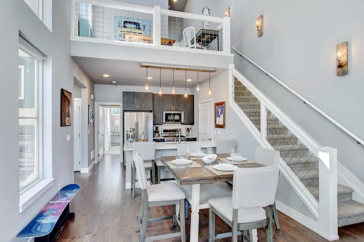 An open-concept space that reveals the dining area, kitchen, and visible loft above. The inviting layout encourages conversation and connection, while the loft area hints at an elevated, private workspace ideal for mixing productivity with relaxation. The scene is bathed in soft, natural light, and the furnishings evoke a sense of mountain-chic decor.