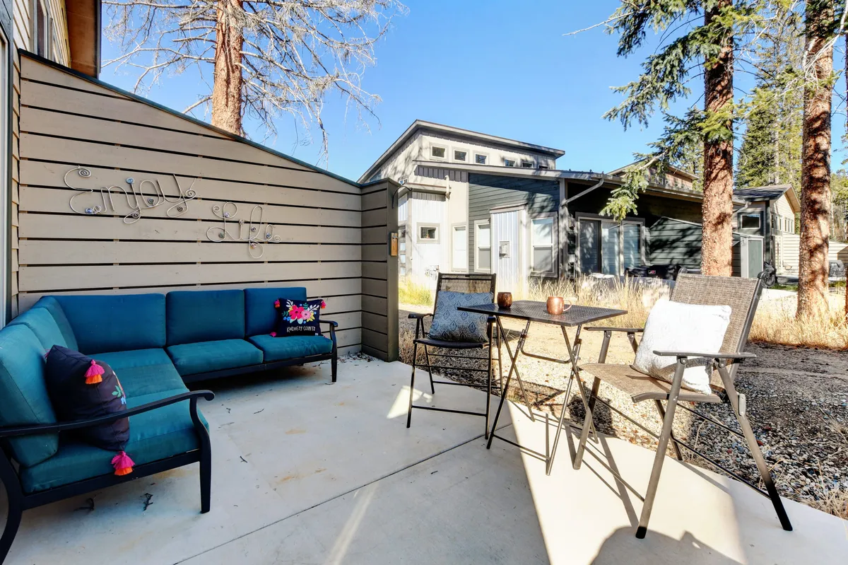 A cozy outdoor seating area featuring a blue couch, a small table with chairs, and decorative pillows set against a wooden wall with text. The space is casual and comfortable and exudes a welcoming vibe, perfect for relaxing or socializing. The patio setting opens up easy access to the outdoors, ideal for guests traveling with pets.