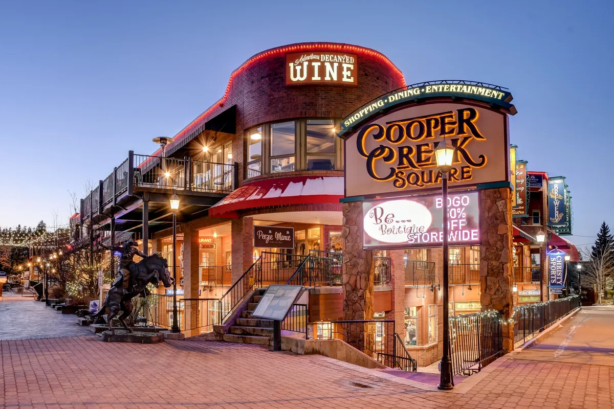 An inviting exterior image of Cooper Creek Square, featuring a lively outdoor social area with a wine shop, seating, and an artistic sculpture. The mood is festive and welcoming, perfect for evening gatherings after a day of adventure.
