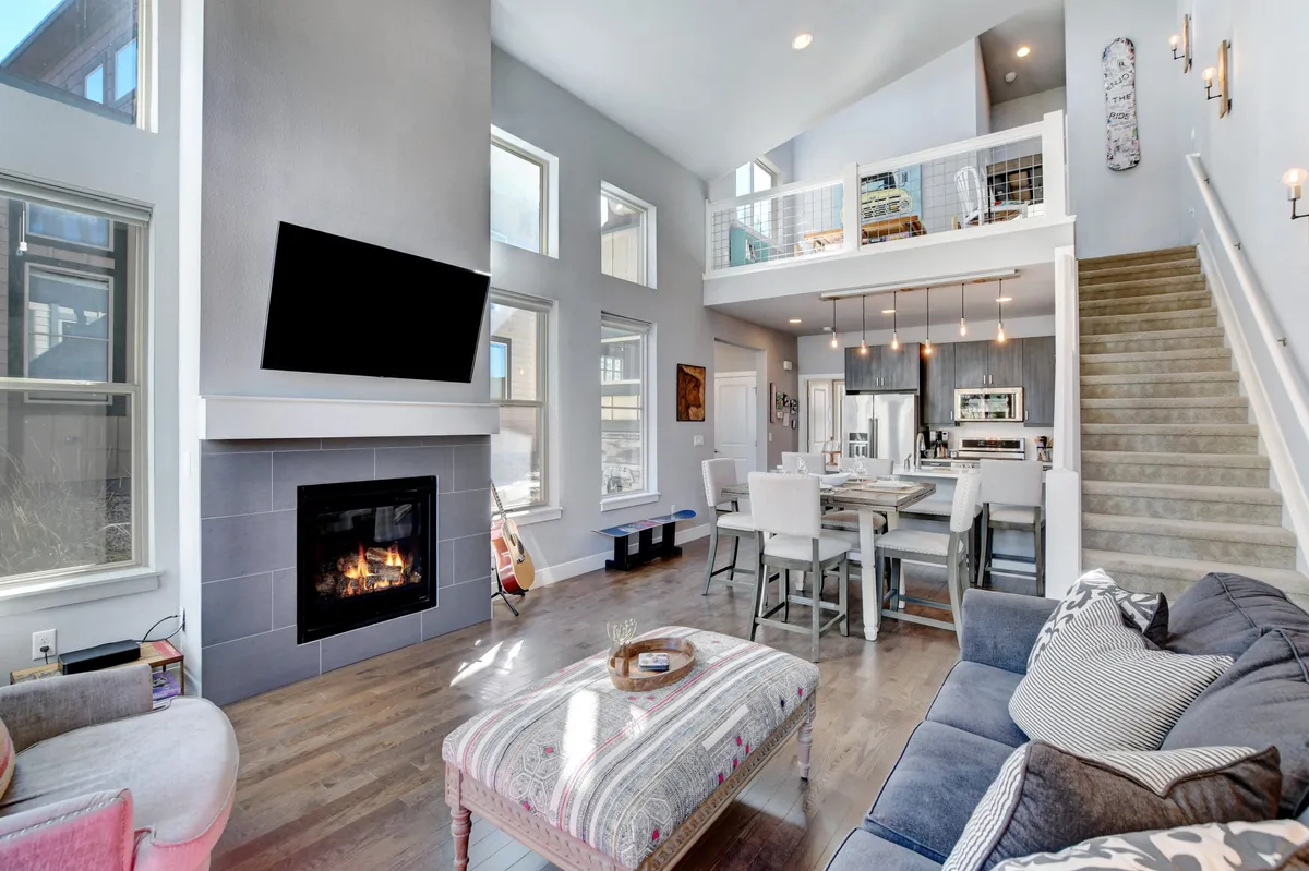The hero image showcases a stylish mountain cabin interior with a crackling fireplace as the centerpiece, a spacious dining area set for gathering, a wooden staircase leading to the second floor, and expansive windows framing scenic mountain views. The setting exudes warmth and comfort, perfect for relaxing or sharing meals after a day of adventure.