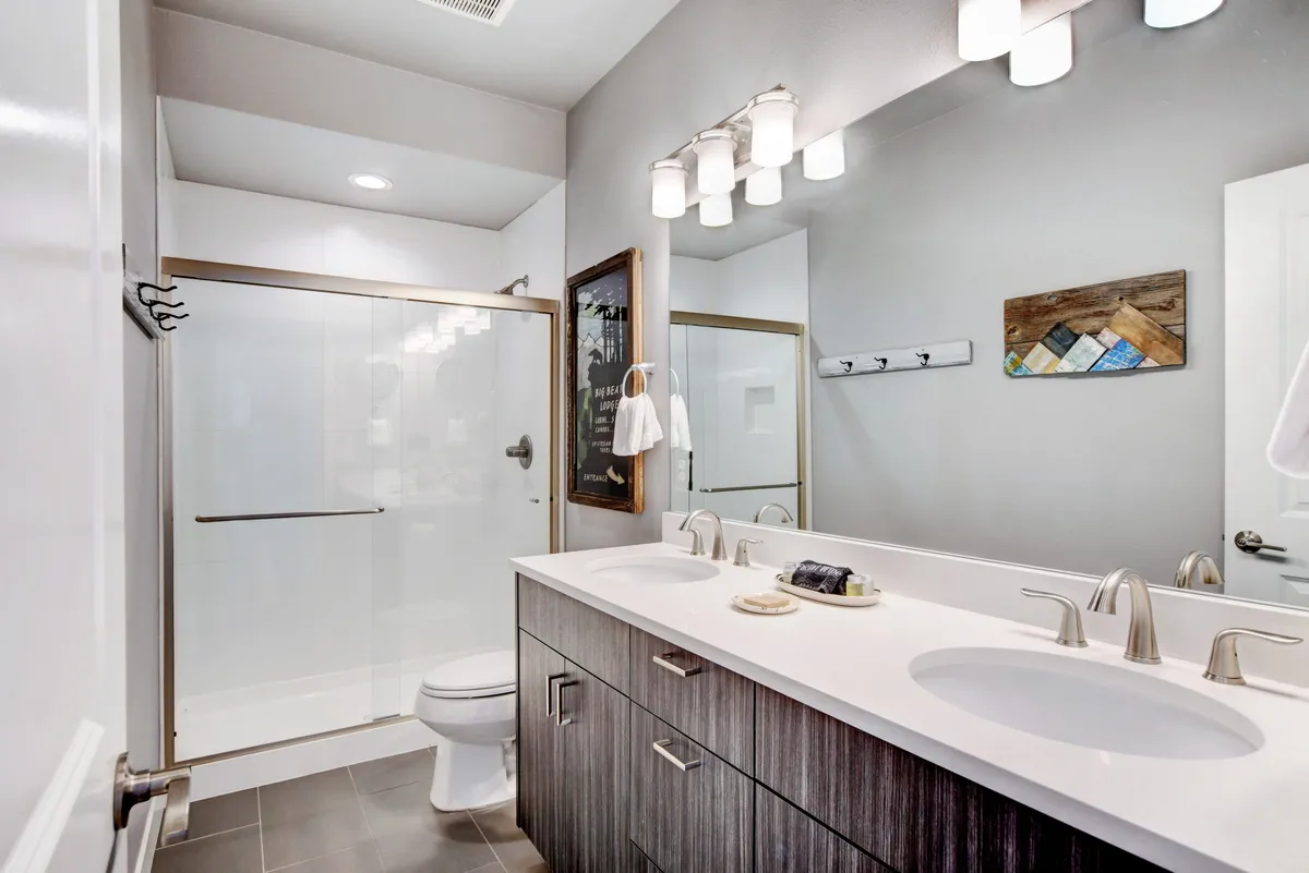 A modern bathroom with a double sink vanity, glass shower enclosure, and framed wall art. Sunlight streams through a window, creating a bright and inviting space ideal for morning routines or winding down after a day outdoors.