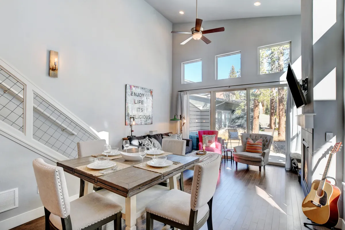 A sunlit dining nook with a table set for a meal, surrounded by colorful accent chairs and large windows that let in ample natural light. A guitar in the corner hints at relaxed, joyful gatherings. The mood is friendly and inviting, perfect for family meals or casual planning sessions.